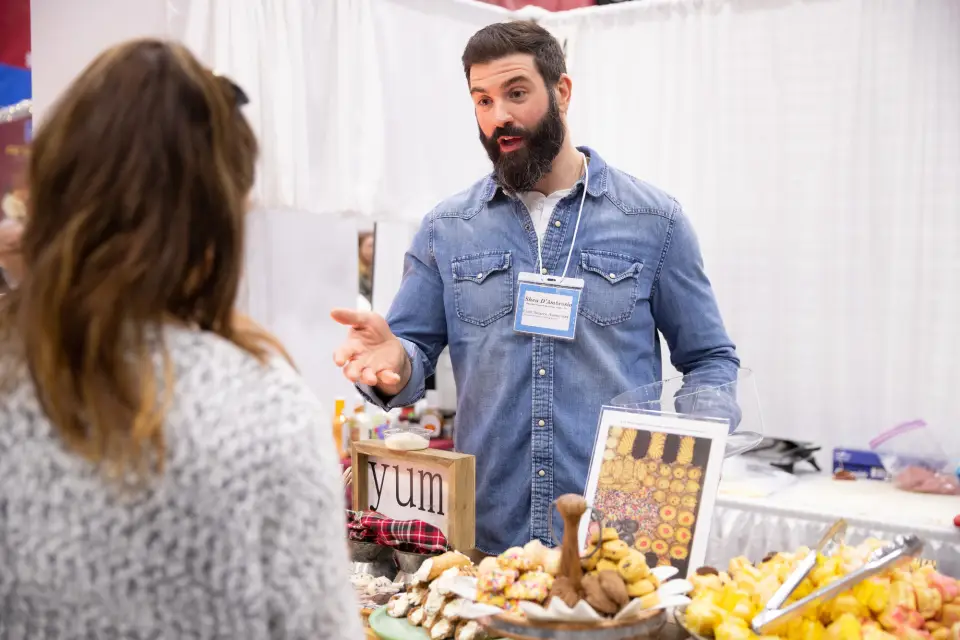 A man explains the table of food in front of him at a convention.