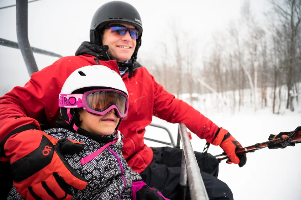 Father has arm around young daughter on ski lift, both wearing ski gear and smiling gently