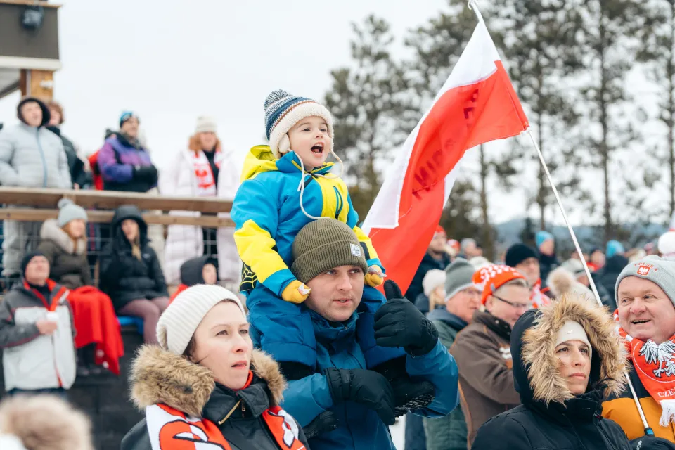 A small child sits on the shoulders of an adult male, in a crowd cheering at a winter outdoor sports event.