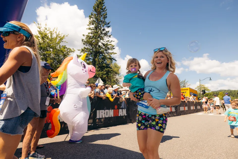 Woman in tank top and shorts holding baby walks past person in unicorn costume at race