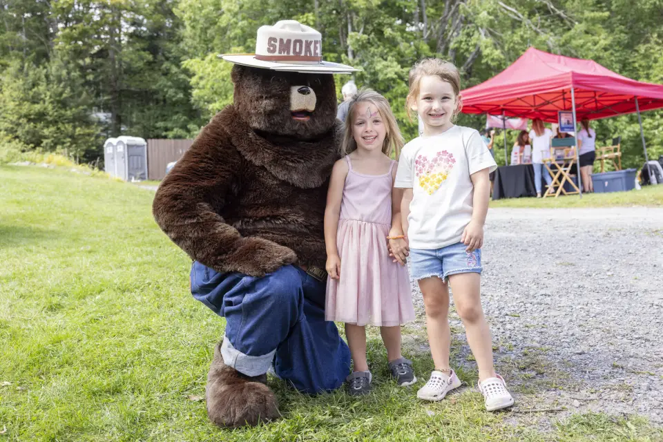 Two children stand next to Smokey Bear, who crouches on the ground next to them.