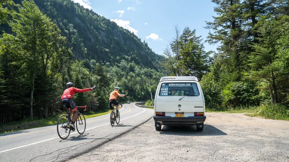 Two cyclists ride single file on shoulder of road  waving to white van parked in pull-off next to them with cliffs and green trees lining the road