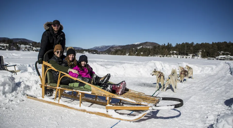 A family poses on a dog sled, with a driver standing on the back of the sled, on a frozen lake.