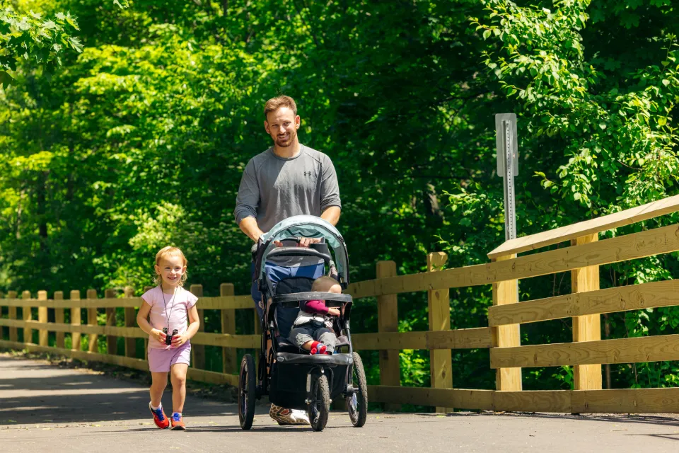 Smiling man pushes stroller with baby while smiling little girl walks beside him holding binoculars on paved section of Rail Trail in summer