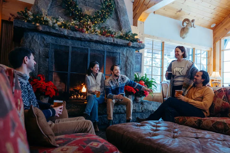 Group of friends sits and stands near fireplace in great room of Grandview Lake Placid with beverages