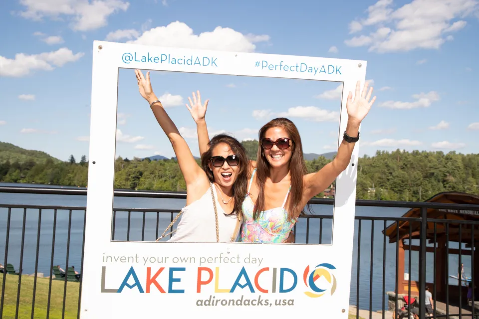 Two women laugh while taking a photo in a Lake Placid frame. 