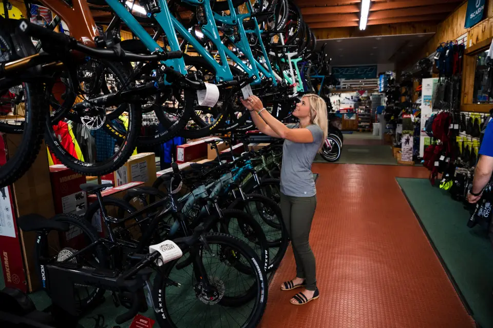 A woman reaches up to a mountain bike on a rack in a cycling shop crowded with bikes.