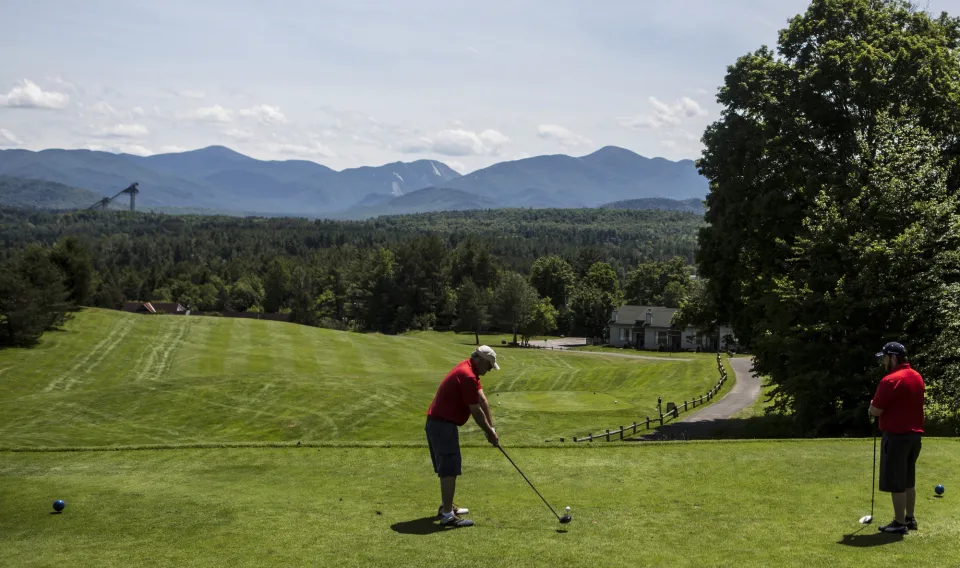 A golfer tees off at a hole looking downhill at a sloping, luxurious golf course with mountains in the distance.