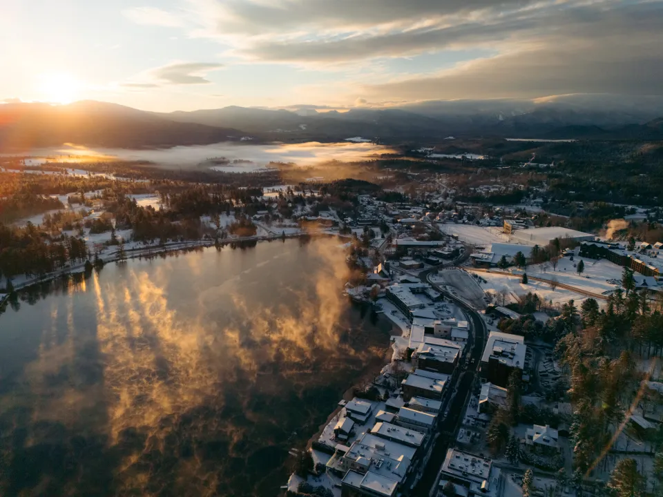 A scenic view of Lake Placid's Main Street and Mirror Lake at sunrise, with mountains in the distance.