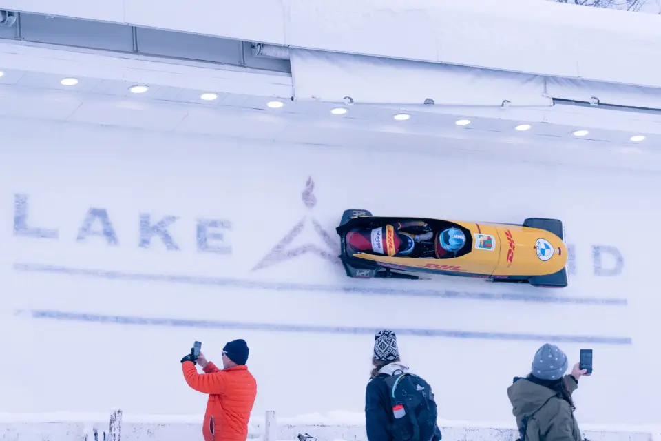 A bobsled rides high on a curve on the Lake Placid bobsled track while spectators watch and take photos.