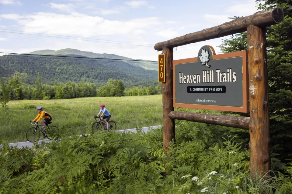 Two cyclists drive by a sign for Heaven Hil Trails in a valley. 