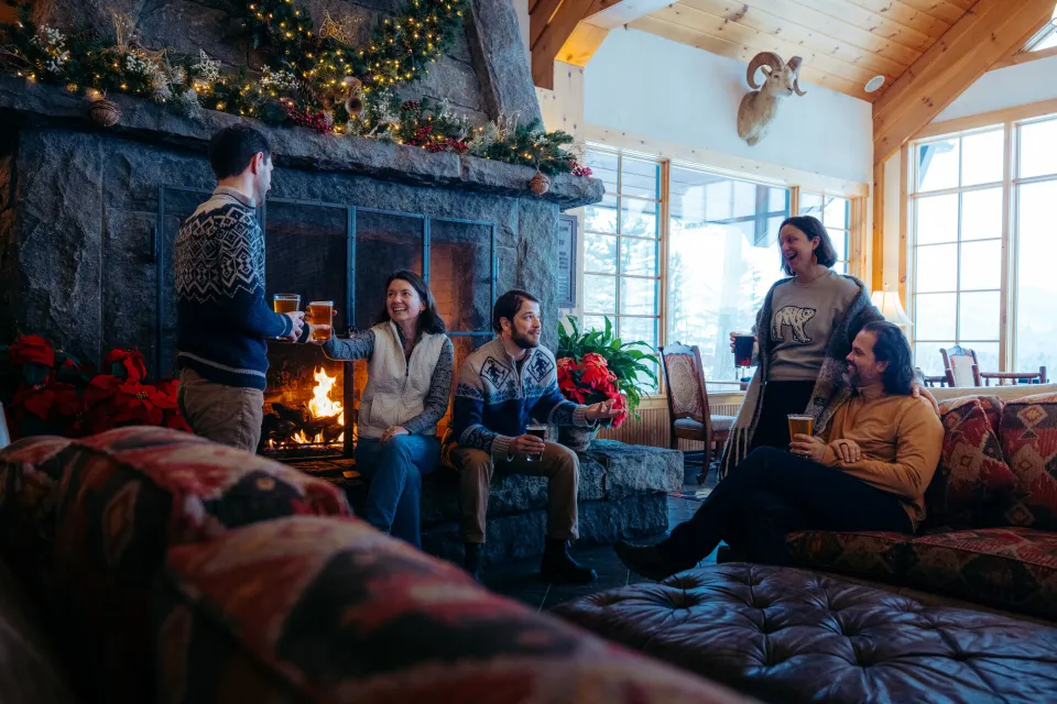A group of friends sit around a grand fireplace in a lodge.