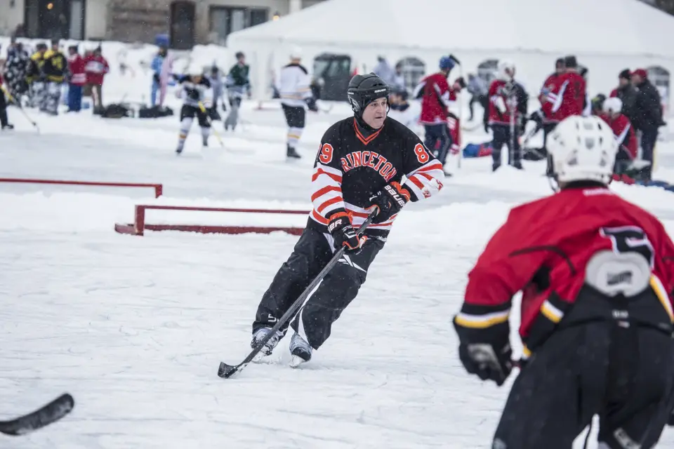 A man in a black hockey jersey with white and orange trim skates on a pond hockey rink.