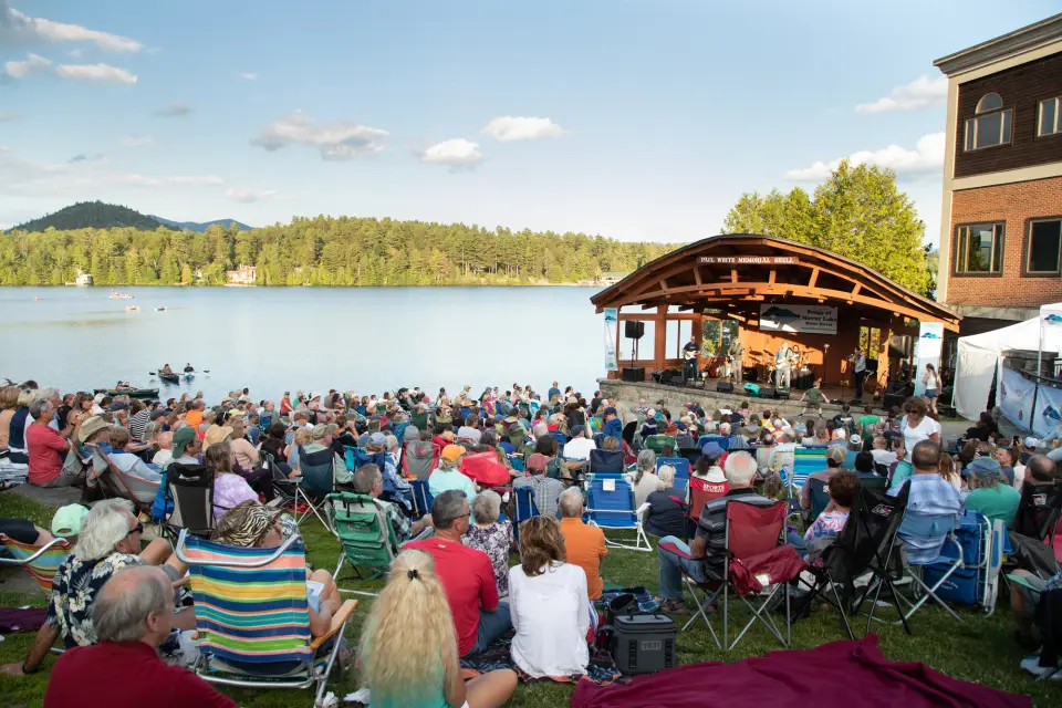 Crowd of people listening to music on lawn in front of bandshell next to lake with mountain views