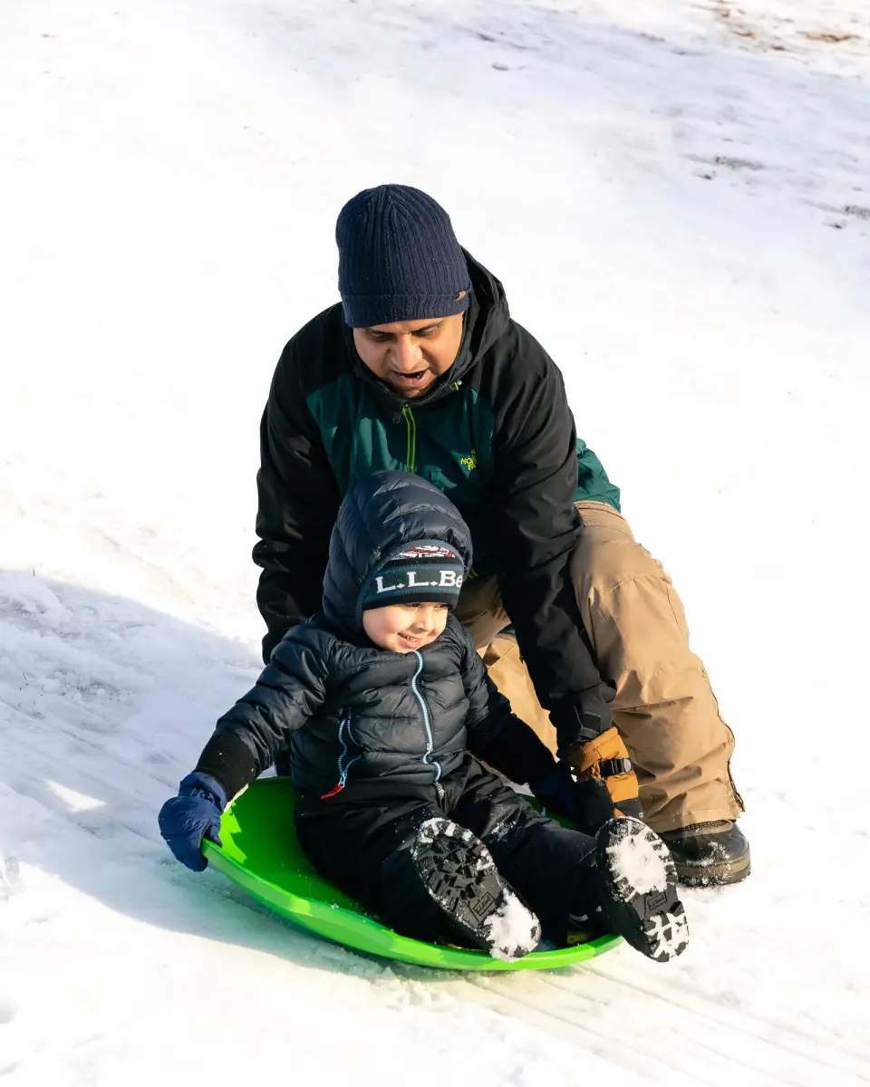 A man in winter outdoor clothing sits on snow behind a little boy on a sledding saucer.