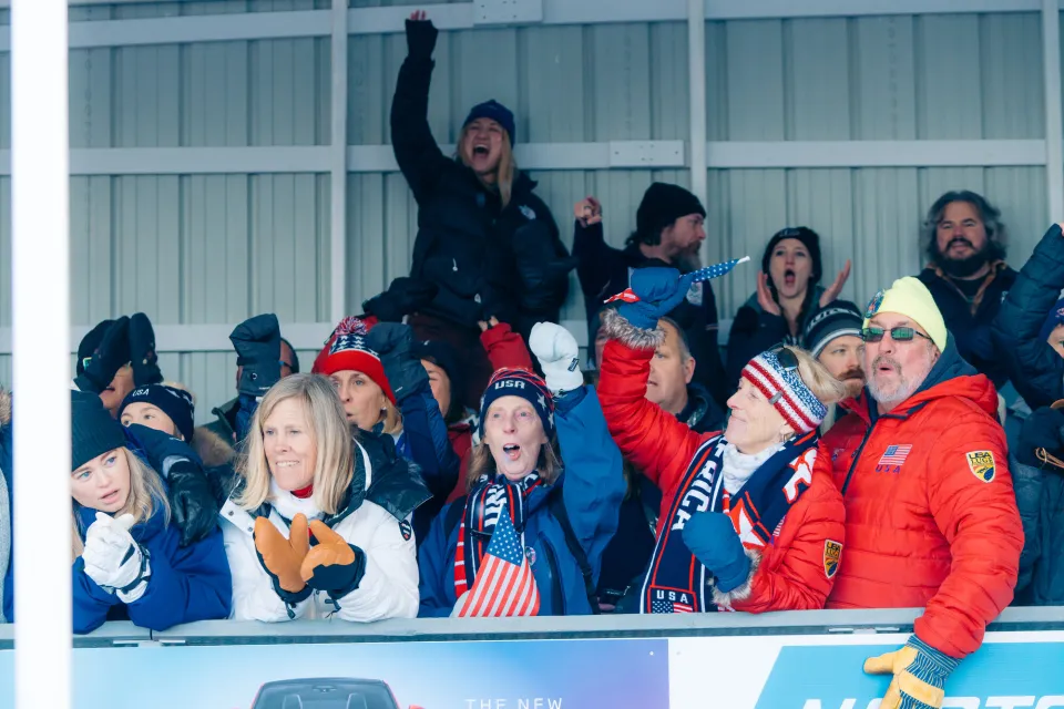 Crowd of people wearing outdoor clothing, many sporting Team USA gear and flags, cheer at the Mt Van Hoevenberg sliding track.