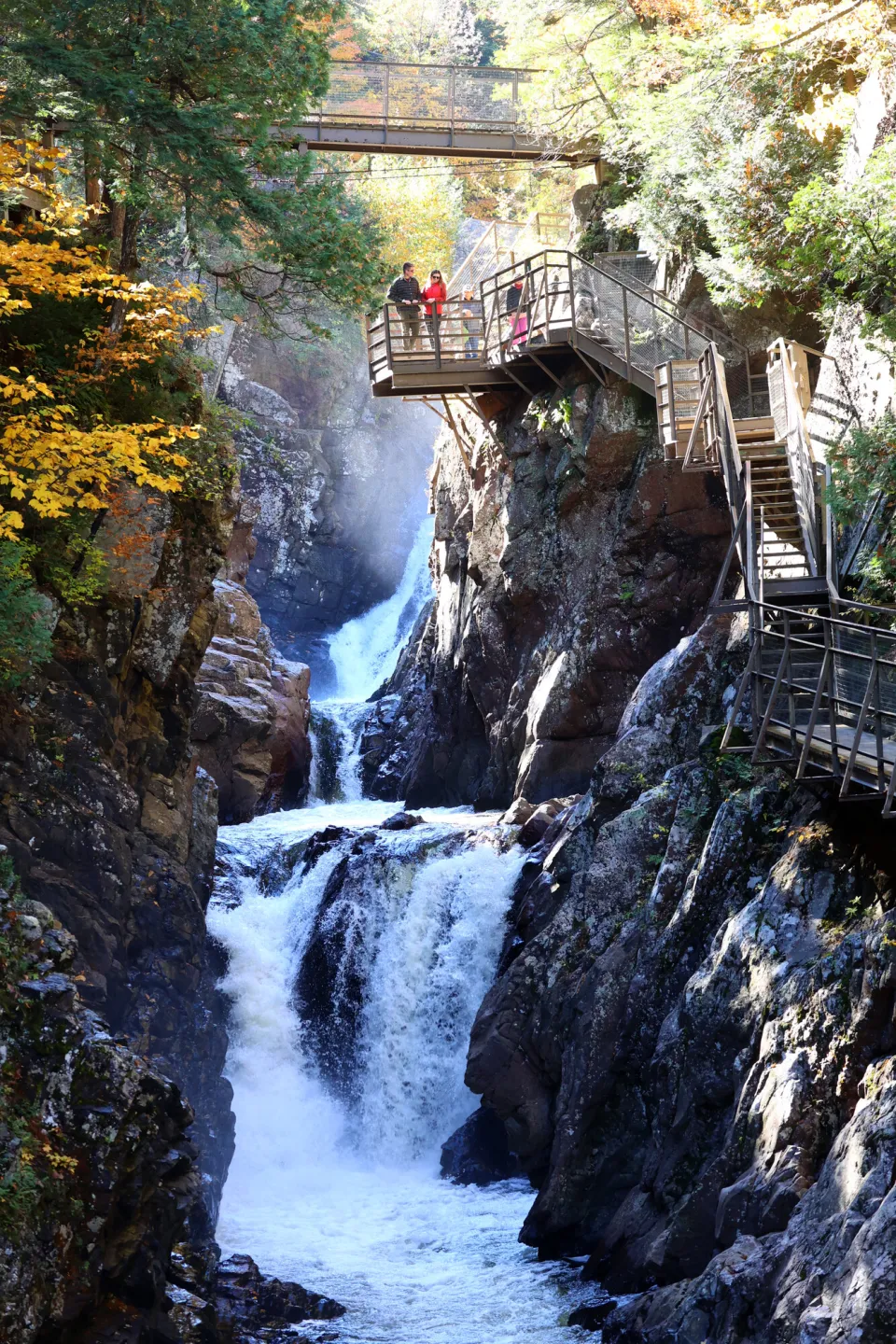 A man and woman stand over a rocky waterfall from a built-in trail alongside it. fall foliage surrounds the waterfall. 
