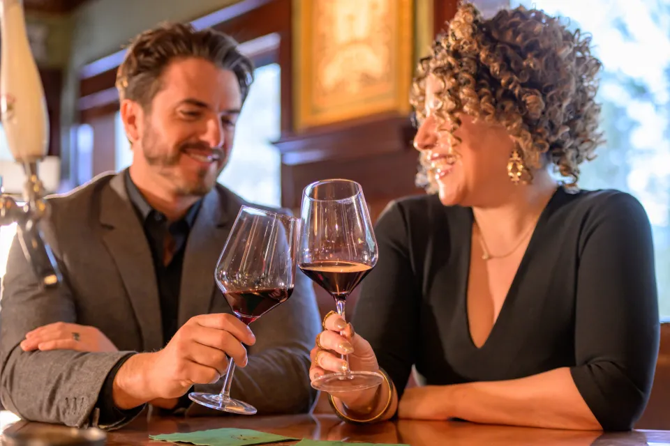 A man and a woman in business casual attire clink glasses of red wine together in a warm-hued fine dining restaurant.
