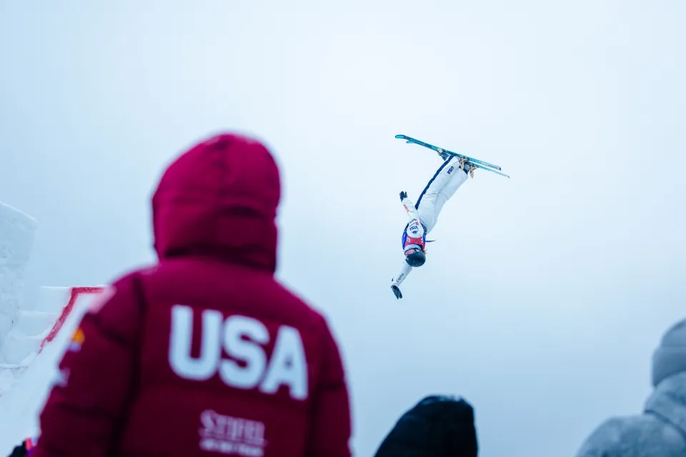 Person in red USA winter coat with hood pulled up stands with back to camera watching aerial ski jumper performing an upside down maneuver