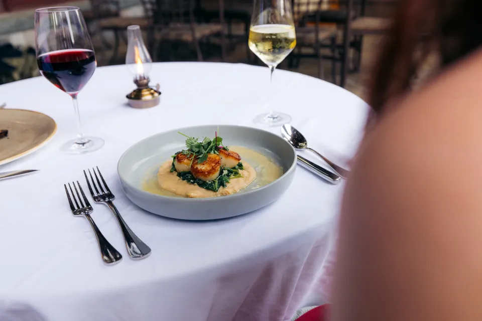 Close-up of a plate of fine dining food on a white plate, with wine glasses, on a table covered with a white tablecloth.