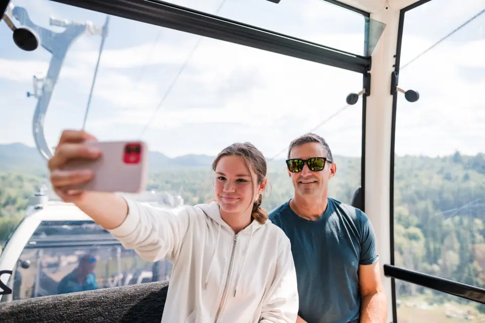 A man and a teenage girl take a selfie inside a glass-enclosed gondola with mountain views behind them.