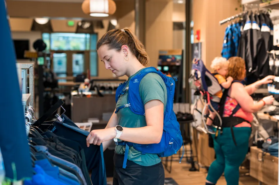 A young woman wearing a hiking water pack browses a rack in an outdoor gear outfitters.