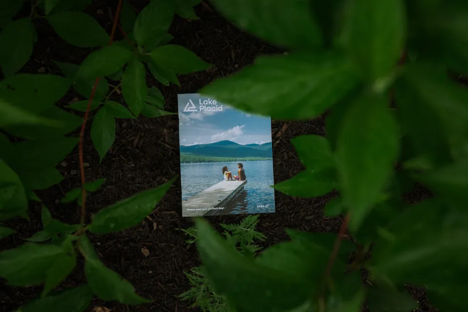 Lake Placid Travel Guide for 2026-27 sits atop dirt and mulch under the cover of green leaves