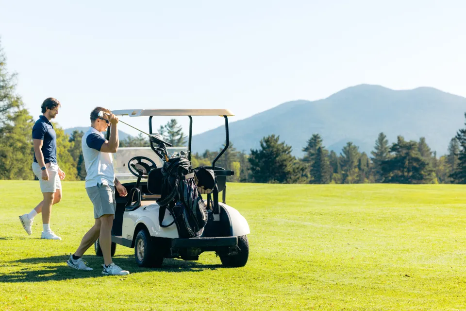 two men get their golf clubs from a golf cart while on a golf course in the mountains.