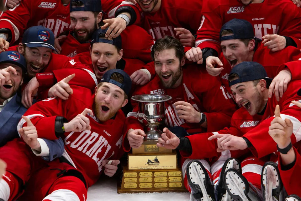 Group of male hockey players crowded around championship trophy on ice wearing red uniforms