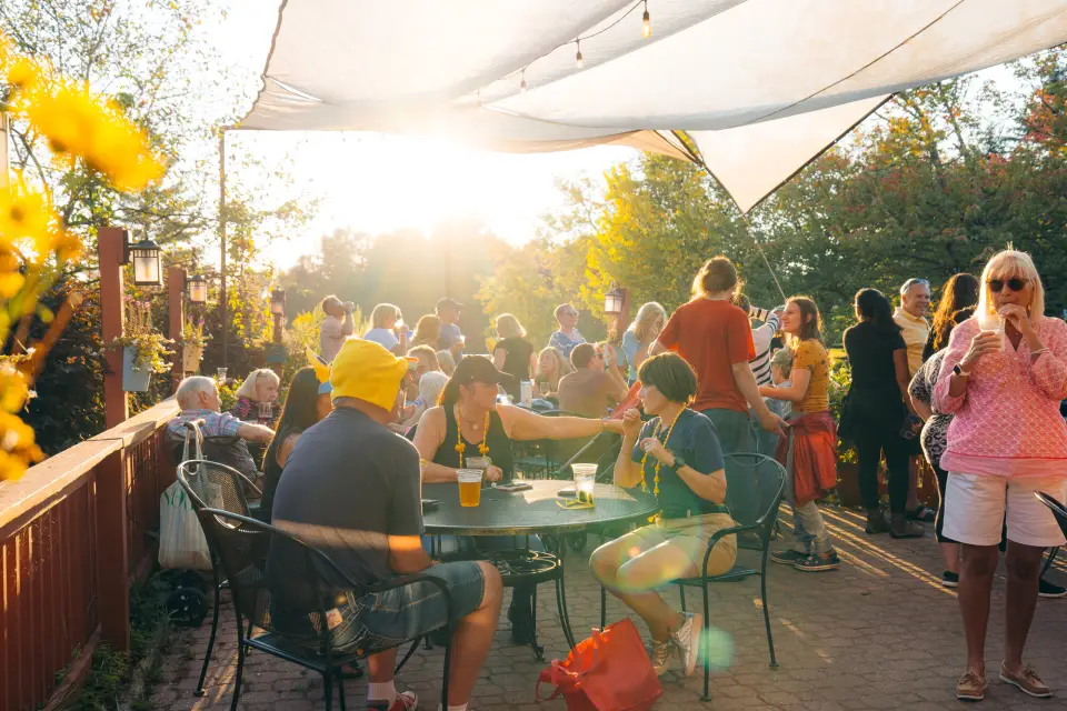 Large group of people on outdoor deck under canvas cover, some sitting at outdoor tables and some standing, mingling at sunset