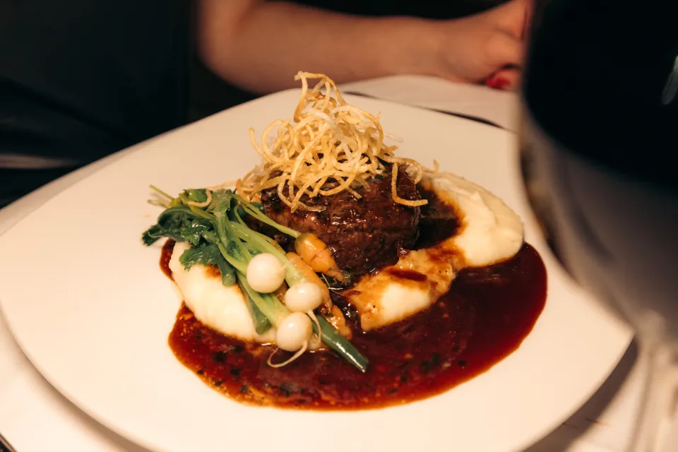 A plate of steak medallion on a bed of greens, mashed potatoes, and sauce. 