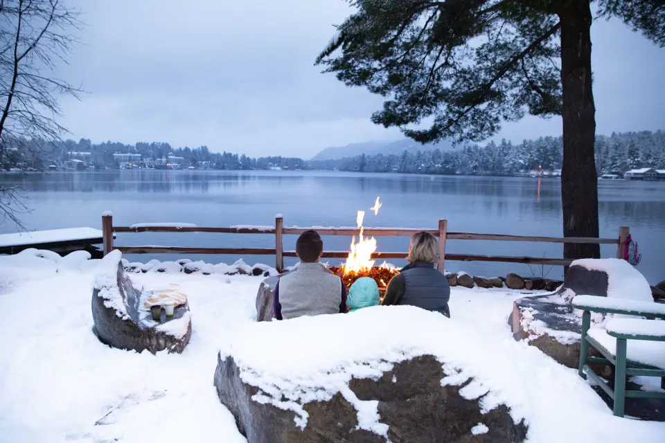 A couple sitting in front of a fire, looking out at Mirror Lake during their stay in Lake Placid.