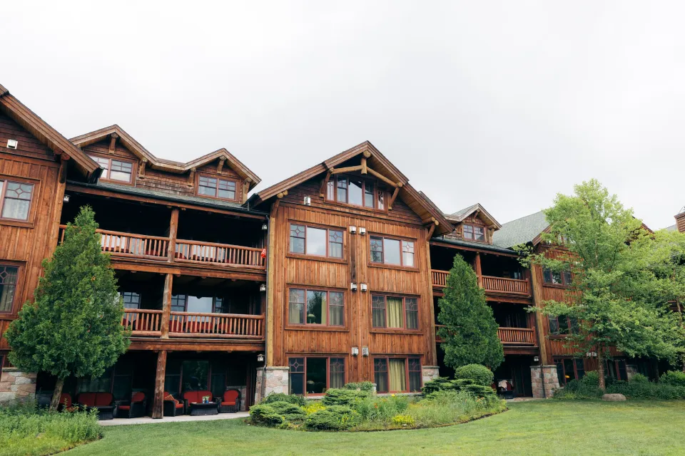 The back of a grand Adirondack lodge with rows of balconies from hotel rooms. Perched over a lush green lawn. 