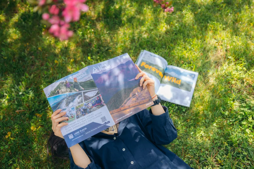 Person lies on their back in green grass under pink flowers reading one Lake Placid Guide with another open next to them on the grass to the Forever Wild page. The guide covers the person's face.