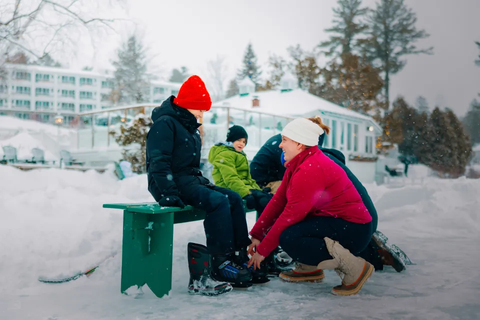 Woman helps a child sitting on a green bench put on ice skates on a frozen lake