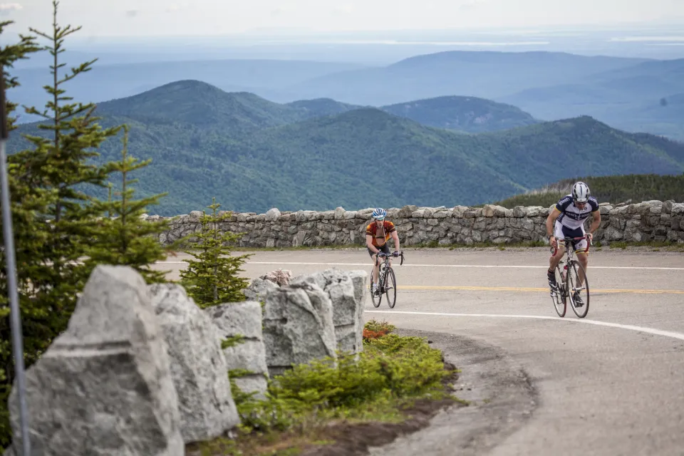 Two cyclists ride up the Whiteface Veterans' Memorial Highway in summer, biking around a turn with stone walls and mountain views