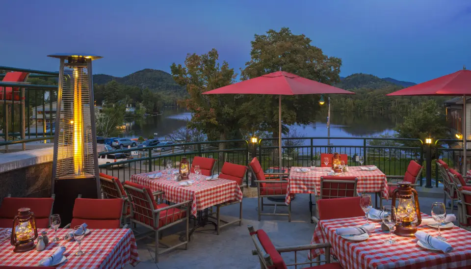 A restaurant deck in early evening dotted with inviting tables, red umbrellas, and a mountain view.
