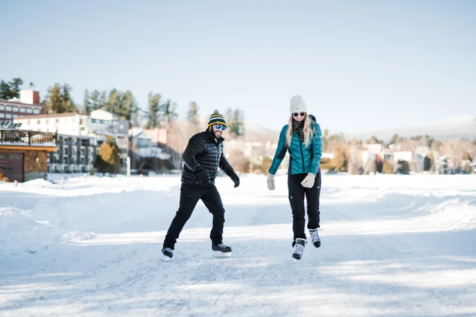 Man and woman in winter outdoor clothing wearing hockey skates skate on shoveled track on Mirror Lake with Lake Placid Main Street buildings in background