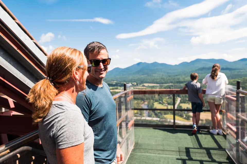 A man and woman smile to each other while their children overlook Olympic ski jumps. 