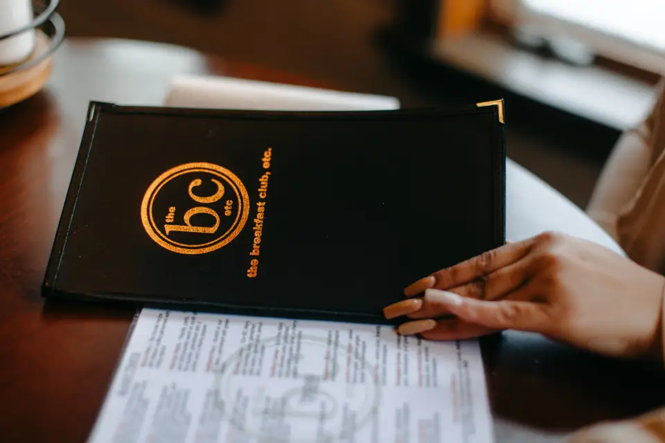 Hand with manicured nails opens Breakfast Club, etc menu with another menu on table