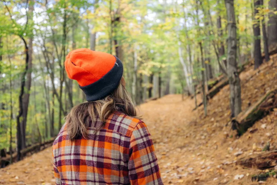 A woman looks away from the camera on a fall trail. She wears an orange beanie and an orange flannel. 