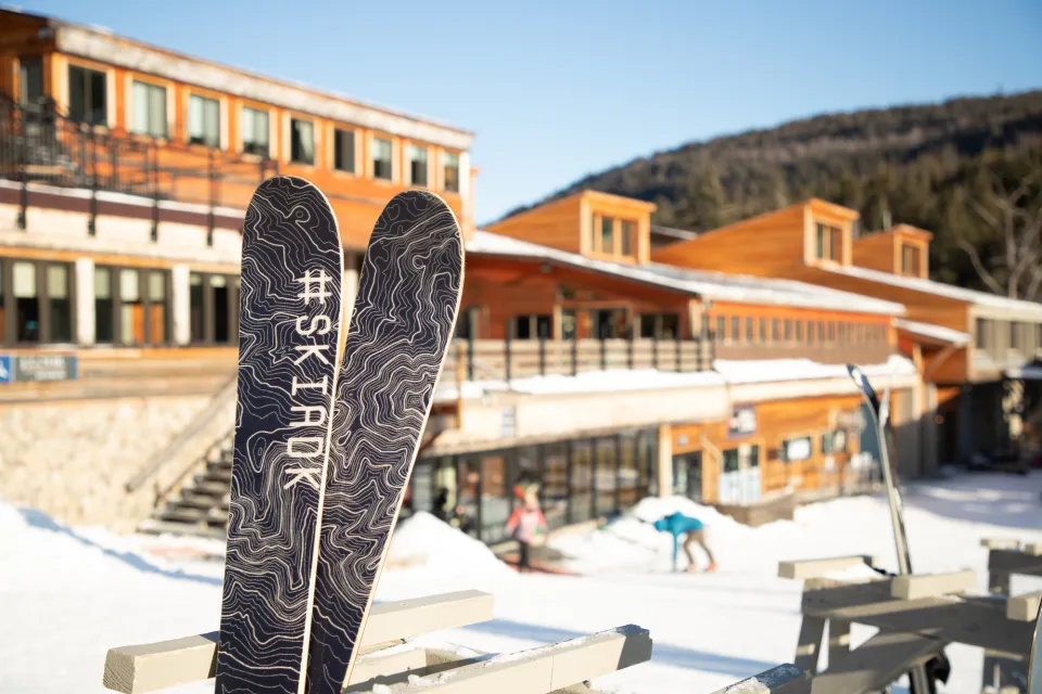 A pair of skis with #ski ADK on them in front of  a grand ski lodge during the day. 