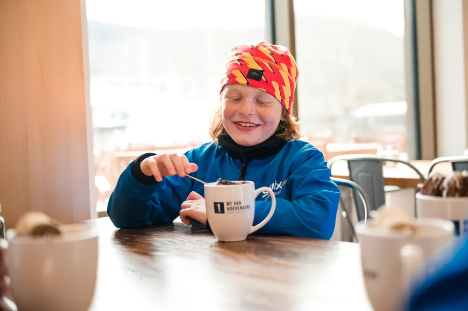 Child plays with wooden marshmallow stick in cup of cocoa wearing winter gear